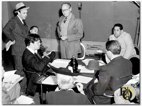 Orson Welles sits at a table, smoking his pipe, with his star, comedian Jack Benny, standing, center, and others during rehearsal of "June Moon" at the KNX radio station for the CBS Campbell Playhouse broadcast, Hollywood, California, March 24. 1940. From left: Bill Morrow (standing); Welles; Benny Rubin sitting behind Welles); Benny; Ernest Chappell (seated at table across from Welles); Jack Beloin (sitting facing camera, another Benny writer). 