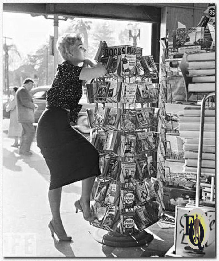 Kim Novak poseert voor een boekenstandaard waarin de Pocket Book uitgave van Ellery Queen's "The Virgin Heiresses" (1954) te koop wordt aangeboden (middenste rij, derde van onderaan). Klik voor een grotere versie.