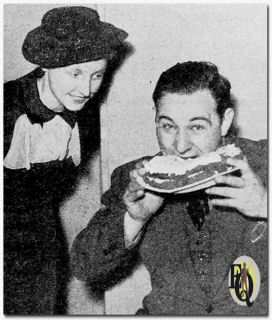 Teddy Bergman trad op als jurylid van de Home State Food Competition tijdens de Annual Women's Competition of Arts and Industries. Hij lijkt te genieten van de prijswinnende Devil's Food cake, terwijl Olive Murphy toekijkt. ("RadioStars", jan 1936)